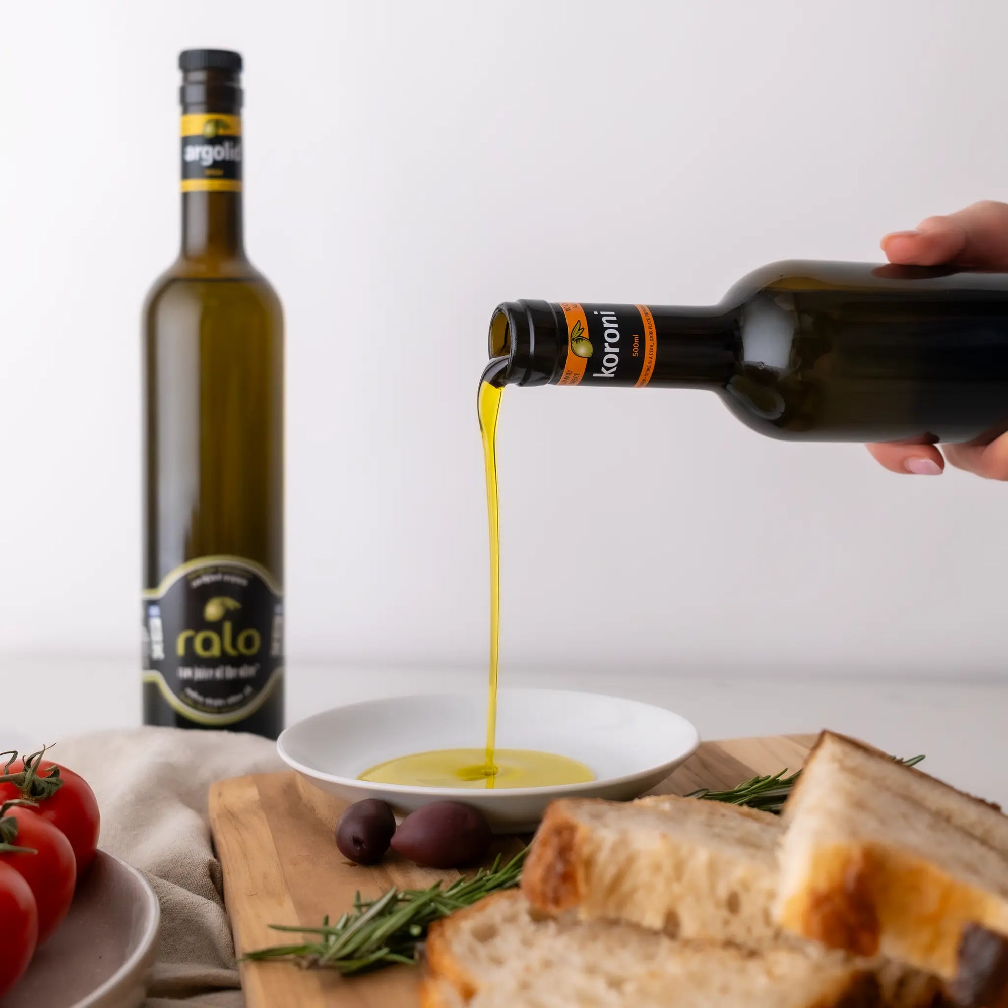 Olive oil being poured from a bottle into a bowl with bread and tomatoes on a wooden board.