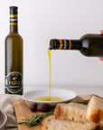 Olive oil being poured from a bottle into a bowl with bread and tomatoes on a wooden board.