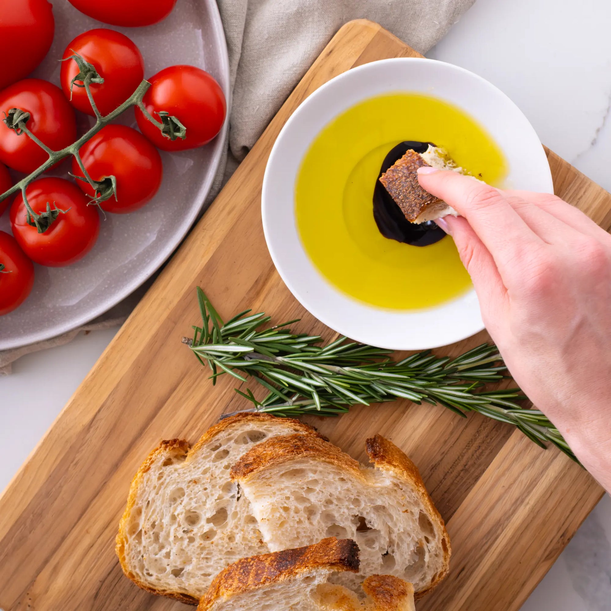 Person dipping bread into olive oil on a wooden board with tomatoes and rosemary.