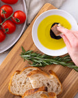 Person dipping bread into olive oil on a wooden board with tomatoes and rosemary.
