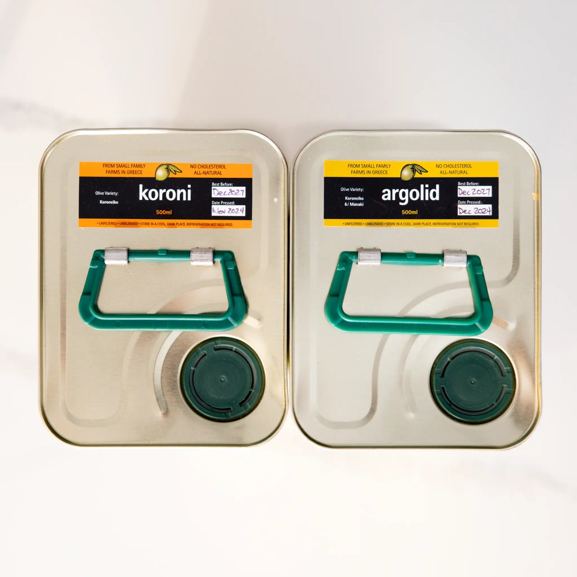 Two metal olive oil tins with green handles, pouring spouts and 'koroni' and 'argolid' stickers on a white background.